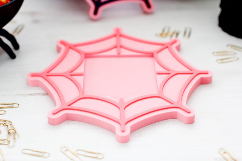 May include: A pink spiderweb-shaped coaster with a square center. The coaster has a raised, darker pink outline and web details. Gold paperclips are scattered around the coaster, suggesting a Halloween or spooky theme.