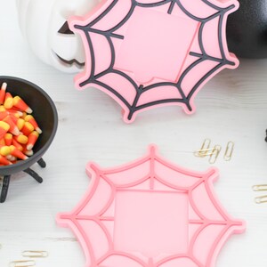 May include: Two pink spiderweb-shaped coasters with black outlines, perfect for Halloween. A white pumpkin with a carved face and a black cauldron are in the background. Candy corn and gold paper clips are also visible.