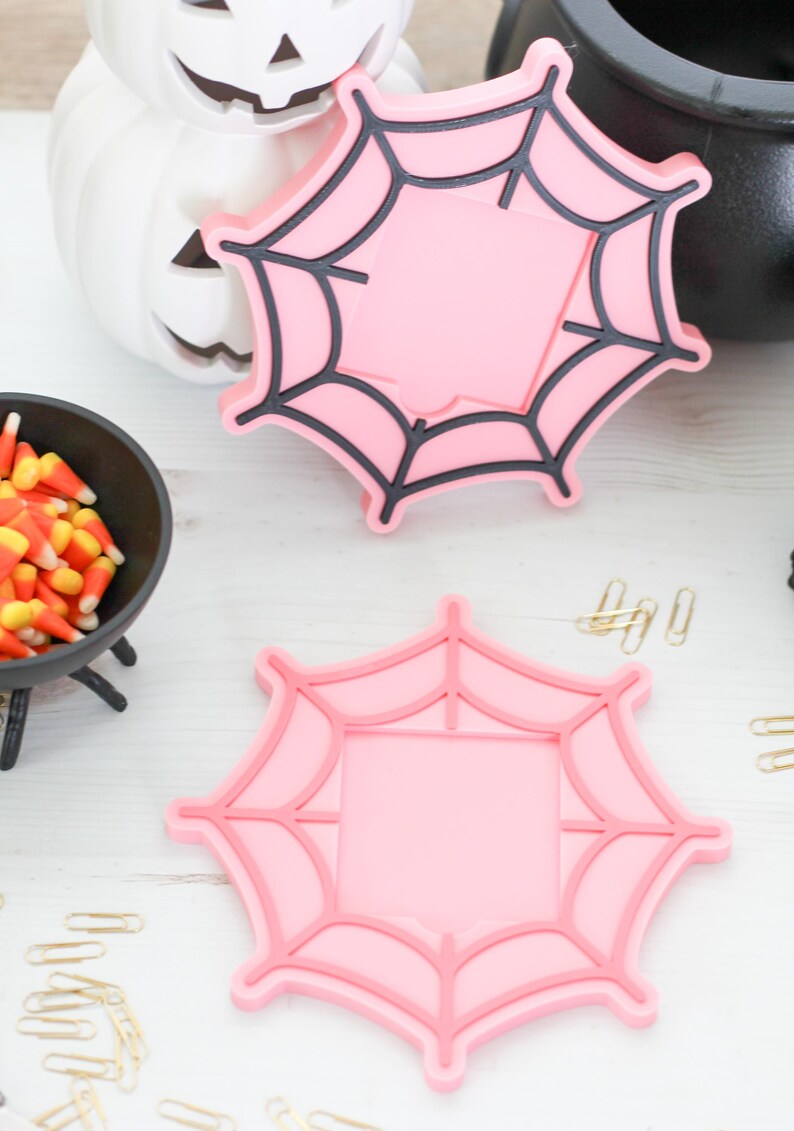 May include: Two pink spiderweb-shaped coasters with black outlines, perfect for Halloween. A white pumpkin with a carved face and a black cauldron are in the background. Candy corn and gold paper clips are also visible.