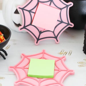 May include: Pink spiderweb-shaped desk organizers with black web detailing, each holding a square sticky note. The Halloween-themed decor includes a white pumpkin, a black cauldron, and candy corn.