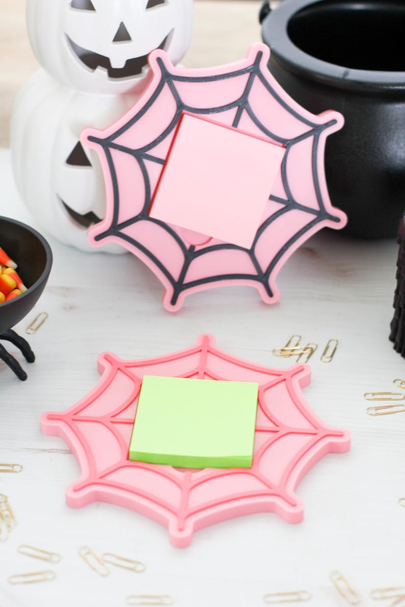 May include: Pink spiderweb-shaped desk organizers with black web detailing, each holding a square sticky note. The Halloween-themed decor includes a white pumpkin, a black cauldron, and candy corn.
