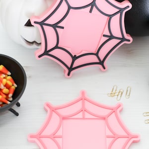 May include: Two pink spiderweb-shaped coasters with black outlines, perfect for Halloween decor. A white pumpkin with a carved face and a black cauldron are in the background. A small bowl of candy corn is also visible.