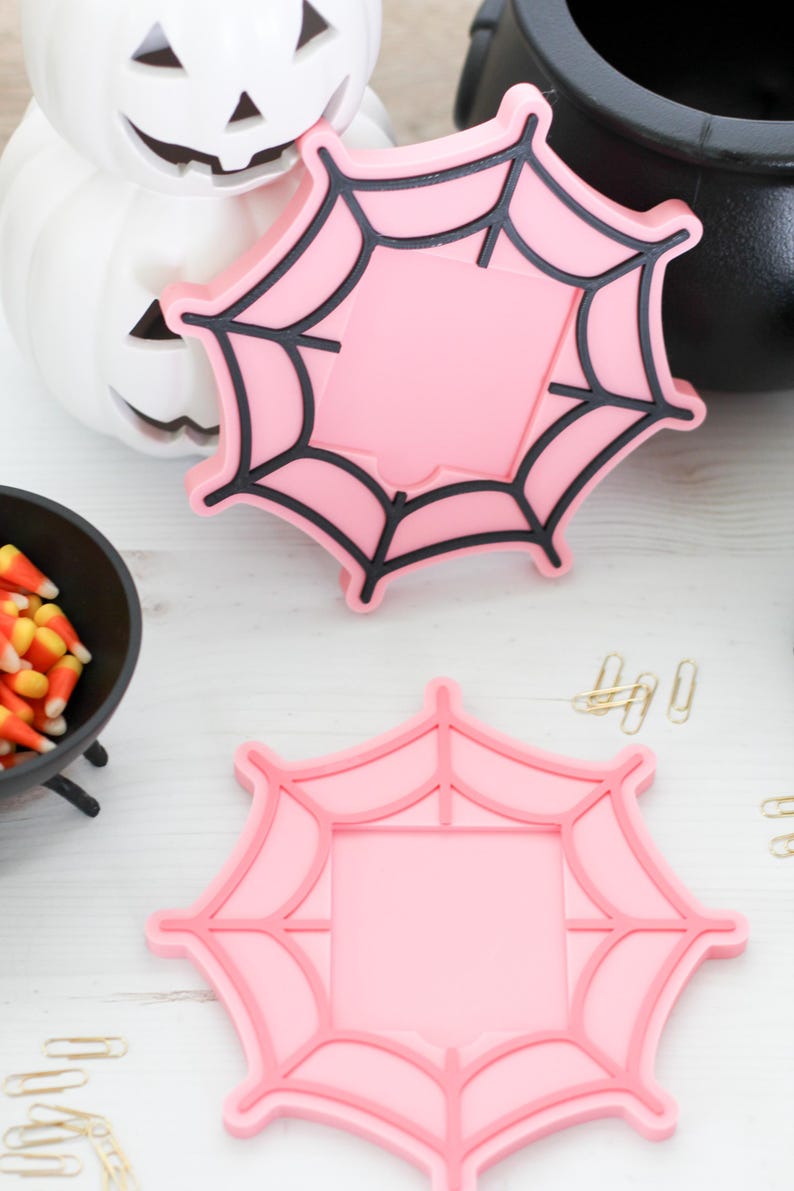 May include: Two pink spiderweb-shaped coasters with black outlines, perfect for Halloween decor. A white pumpkin with a carved face and a black cauldron are in the background. A small bowl of candy corn is also visible.