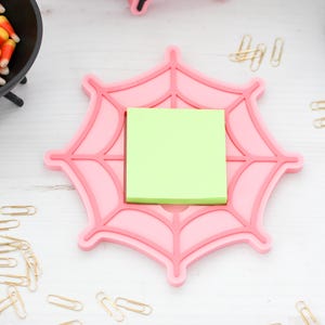 May include: A pink spiderweb-shaped desk organizer with a green sticky note pad. The organizer has a raised design and is surrounded by gold paper clips and a pair of scissors. A small black cauldron with candy corn is in the background.