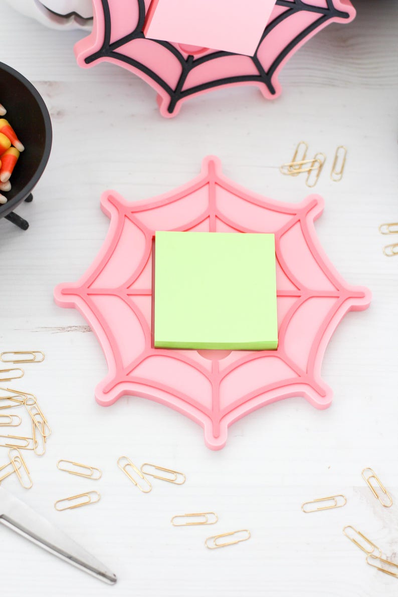 May include: A pink spiderweb-shaped desk organizer with a green sticky note pad. The organizer has a raised design and is surrounded by gold paper clips and a pair of scissors. A small black cauldron with candy corn is in the background.