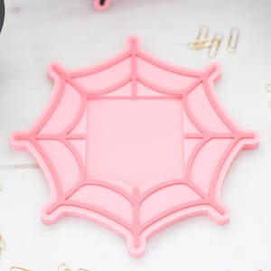 May include: A pink spiderweb-shaped cookie cutter with a square center. The cutter is on a white surface, with candy corn and paper clips nearby. This item is perfect for Halloween-themed baking.