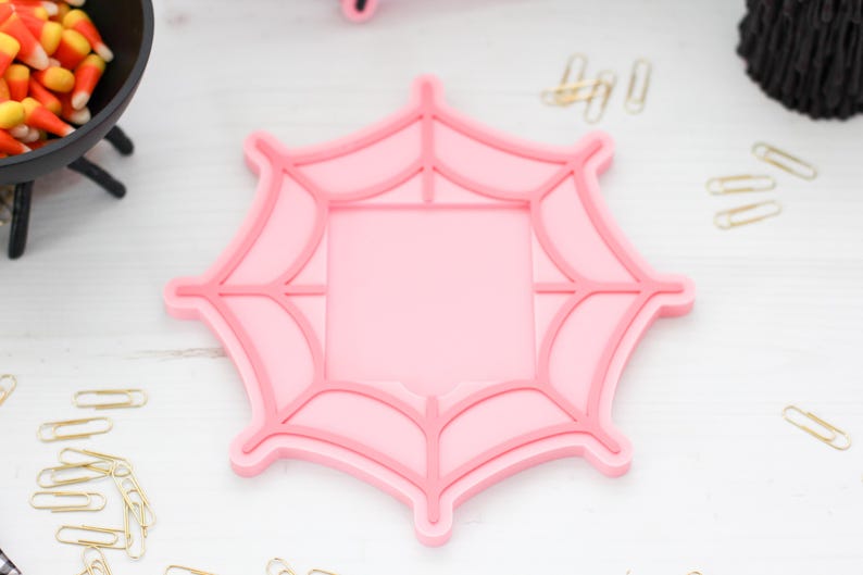 May include: A pink spiderweb-shaped cookie cutter with a square center. The cutter is on a white surface, with candy corn and paper clips nearby. This item is perfect for Halloween-themed baking.
