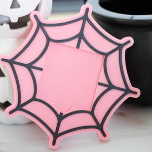 May include: A pink spiderweb-shaped coaster with black outlines, perfect for Halloween decor. The coaster is set against a backdrop of a white pumpkin, a black cauldron, and a black candle, creating a spooky theme.