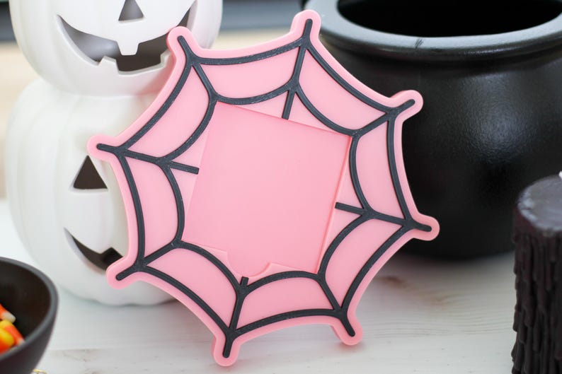 May include: A pink spiderweb-shaped coaster with black outlines, perfect for Halloween decor. The coaster is set against a backdrop of a white pumpkin, a black cauldron, and a black candle, creating a spooky theme.