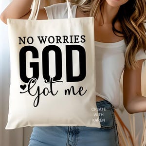 May include: Cream-colored tote bag with the text "NO WORRIES GOD got me" in black. The words "CREATE WITH KAREN" are printed in white at the bottom right corner. The bag has two cream-colored handles.