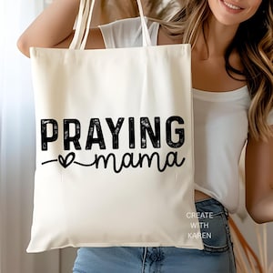 May include: Cream-colored tote bag with the words "PRAYING mama" in black, stylized text. A heart symbol is between the words. The text "CREATE WITH KAREN" is printed in the lower right corner.