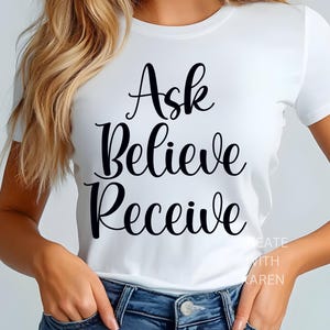 May include: White t-shirt featuring the words "Ask Believe Receive" in black script. The shirt is short-sleeved and paired with blue jeans. The text is in a cursive font. The shirt is a casual style.