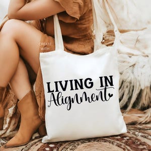 May include: Cream-colored tote bag with the words "LIVING IN Alignment" in black script. The bag has long straps and is made of a soft, likely cotton, material. The words "CREATE WITH KAREN" are printed on the side of the bag.
