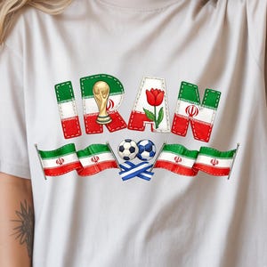 May include: Beige t-shirt with a colorful graphic design. The word "IRAN" is spelled out in a patchwork style with the Iranian flag colors, a trophy, and a tulip. Below are two soccer balls and Iranian flags.