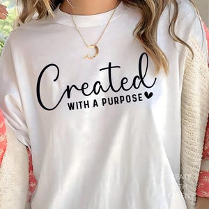 Könnte beinhalten: Weißes T-Shirt mit dem Schriftzug "Created with a Purpose" in schwarzer Schrift. Das Shirt hat einen Rundhalsausschnitt und kurze Ärmel. Eine goldene Halskette ist sichtbar. Das Model trägt eine rosa-weiße Strickjacke.