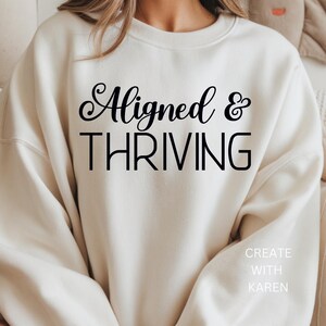 May include: Cream-colored sweatshirt with the words "Aligned & THRIVING" in black script and block letters. The text "CREATE WITH KAREN" is printed below. The sweatshirt has a crew neck and long sleeves.