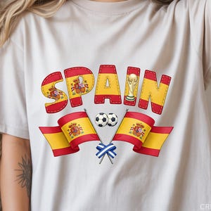Może przedstawiać: Kremowa koszulka z nadrukiem graficznym z napisem "SPAIN" w kolorze czerwonym i żółtym, zawierającym hiszpańskie flagi, piłki nożne i trofeum. Projekt zawiera tekst "CREATE WITH KAREN".