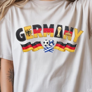 May include: Cream-colored t-shirt with a colorful graphic design featuring the word "GERMANY" in a stitched font. The design includes German flags, soccer balls, and the German coat of arms. The shirt is a casual wear item.