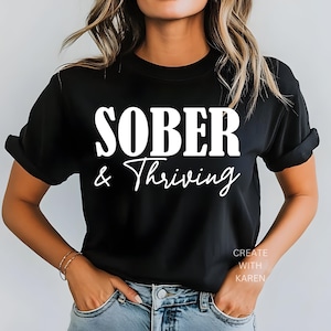 May include: Black t-shirt with the words "SOBER & Thriving" in white block and script font. The shirt is worn by a person in blue jeans. The text "CREATE WITH KAREN" is printed in white on the lower right side of the shirt.