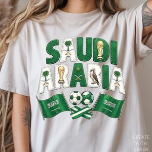 May include: Beige t-shirt with a green and white graphic design that says "SAUDI ARABIA". The design includes a trophy, soccer balls, flags, and a falcon. The text is stitched with green thread.