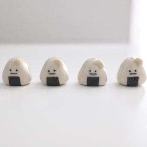 Puede incluir: Cuatro pequeñas figuras de cerámica blancas en forma de onigiri, una bola de arroz japonesa. Cada una tiene una base de algas nori negras y una cara simple. Las figuras están dispuestas en fila sobre una superficie blanca.