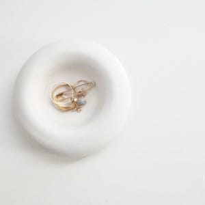 Op de afbeelding: Een witte, cirkelvormige sieradenschaal met een ondiepe kom. In de kom liggen verschillende gouden ringen en een veiligheidsspeld. De schaal staat op een wit oppervlak, wat een minimalistische esthetiek creëert. De sieraden lijken van goud te zijn.