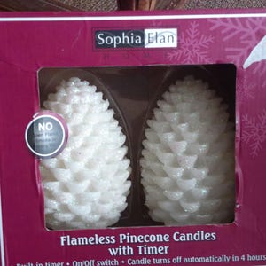 May include: Two white flameless pinecone candles with glitter accents are displayed in a burgundy box. The box has the text "Sophia Elan HOME" and "Flameless Pinecone Candles with Timer". The candles have an on/off switch and turn off automatically in 4 hours.
