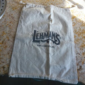 Può includere: Un sacco di tela colorata con coulisse con la scritta "LEHMAN'S FOR A SIMPLER LIFE" in blu scuro. La borsa è rettangolare e si trova su un piano di lavoro maculato.
