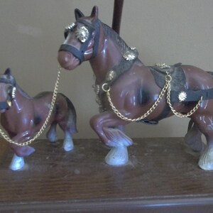 May include: A pair of brown horse figurines with gold-colored chain reins. The larger horse has decorative gold accents on its bridle and harness. The horses stand on a wooden base, creating a decorative display.