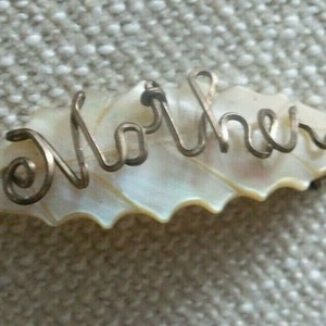 Op de afbeelding: Een witte parelmoer schelp met het woord "Moeder" gespeld in zilverdraad.