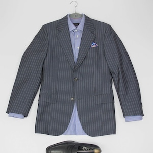 May include: A dark gray pinstripe blazer with a light blue button-down shirt and a small patterned pocket square. Two black leather loafers with a silver inscription are placed below the blazer.
