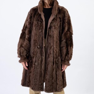 Può includere: Un lungo cappotto di pelliccia marrone con chiusura a zip e bottoni. Il cappotto ha un colletto alto e maniche lunghe.