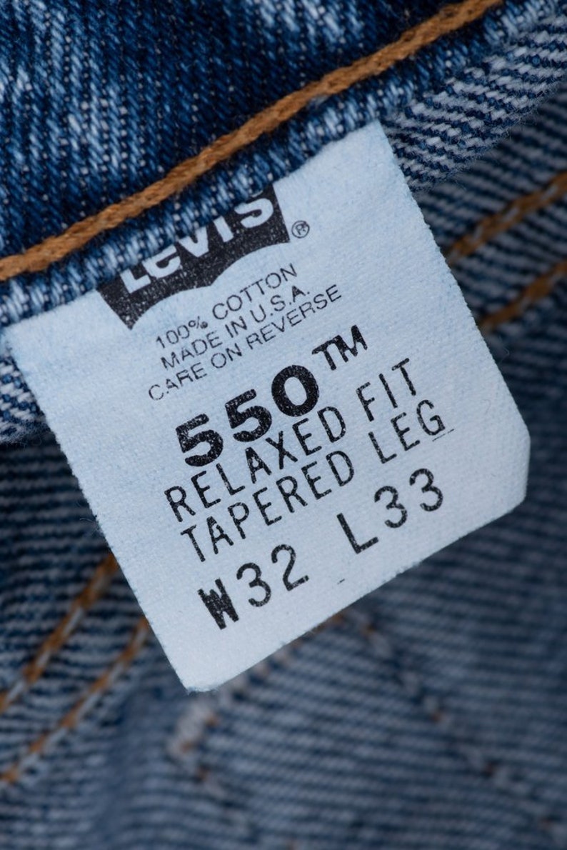 Może przedstawiać: Biała metka dżins&oacute;w Levi's z czarnym tekstem. Metka zawiera napis "550 TM Relaxed Fit Tapered Leg W32 L33". Zawiera r&oacute;wnież napis "100% Bawełna Wyprodukowano w USA. Instrukcja pielęgnacji na odwrocie".