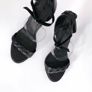 May include: A pair of black leather heeled sandals with braided straps. The sandals have a closed toe and an open heel.