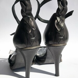 May include: A pair of black leather heeled sandals with braided straps. The sandals have a clear strap across the front and a buckle closure at the ankle.