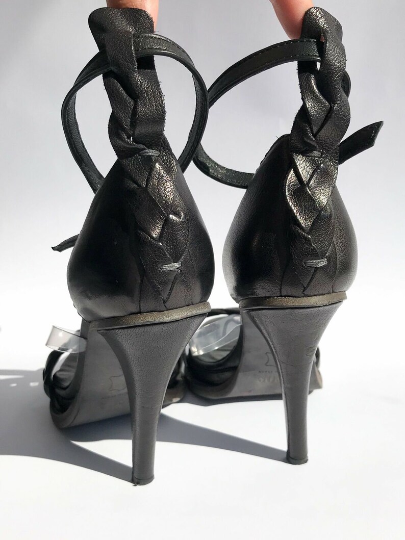 May include: A pair of black leather heeled sandals with braided straps. The sandals have a clear strap across the front and a buckle closure at the ankle.