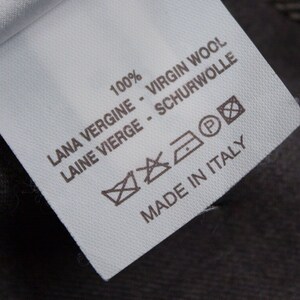 Puede incluir: Etiqueta de cuidado de la tela blanca con texto negro y s&iacute;mbolos. El texto dice "100% VIRGIN WOOL LANA VERGINE - LAINE VIERGE - SCHURWOLLE" y "MADE IN ITALY". Los s&iacute;mbolos incluyen una lavadora, una plancha, un s&iacute;mbolo de limpieza en seco y un c&iacute;rculo con una "P" dentro.