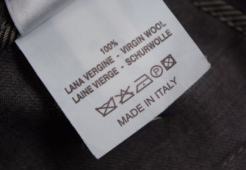 Puede incluir: Etiqueta de cuidado de la tela blanca con texto negro y s&iacute;mbolos. El texto dice "100% VIRGIN WOOL LANA VERGINE - LAINE VIERGE - SCHURWOLLE" y "MADE IN ITALY". Los s&iacute;mbolos incluyen una lavadora, una plancha, un s&iacute;mbolo de limpieza en seco y un c&iacute;rculo con una "P" dentro.