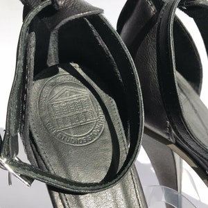 May include: Black leather heeled sandals with an ankle strap and a buckle closure. The sandals have a logo on the insole that says "Cross Stone Studios".