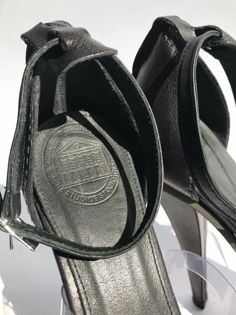 May include: Black leather heeled sandals with an ankle strap and a buckle closure. The sandals have a logo on the insole that says "Cross Stone Studios".