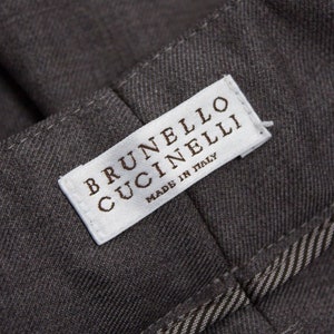 Puede incluir: Una etiqueta blanca con el texto "BRUNELLO CUCINELLI" y "MADE IN ITALY" sobre un fondo de tela gris.
