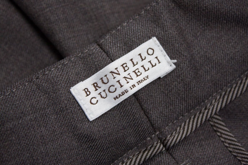 Puede incluir: Una etiqueta blanca con el texto "BRUNELLO CUCINELLI" y "MADE IN ITALY" sobre un fondo de tela gris.