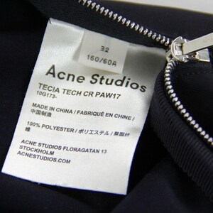 Puede incluir: Primer plano de una prenda azul marino con cremallera plateada y etiqueta blanca. La etiqueta dice "Acne Studios", el texto "TECIA TECH CR PAW17" y "MADE IN CHINA".