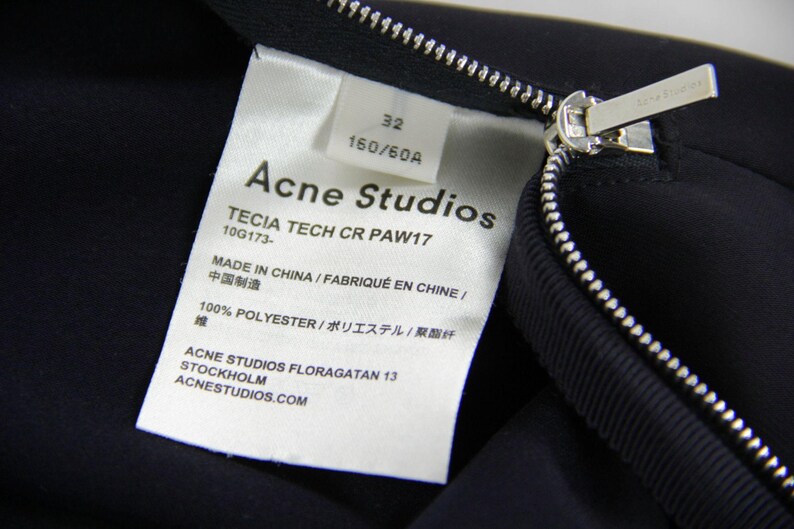Puede incluir: Primer plano de una prenda azul marino con cremallera plateada y etiqueta blanca. La etiqueta dice "Acne Studios", el texto "TECIA TECH CR PAW17" y "MADE IN CHINA".