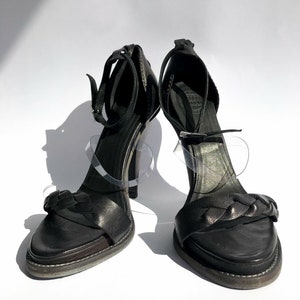 May include: A pair of black leather heeled sandals with a braided strap detail. The sandals have a high heel and an open toe.