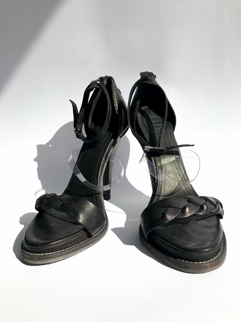May include: A pair of black leather heeled sandals with a braided strap detail. The sandals have a high heel and an open toe.