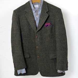 Peut inclure: Un blazer en tweed vert foncé avec un motif en chevrons, trois boutons marron et une pochette en soie à motifs cachemire. Une chemise bleue claire à motifs est visible en dessous. Le blazer est suspendu à un cintre en bois.