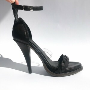 May include: Black leather heeled sandals with a braided strap and an ankle strap with a silver buckle.