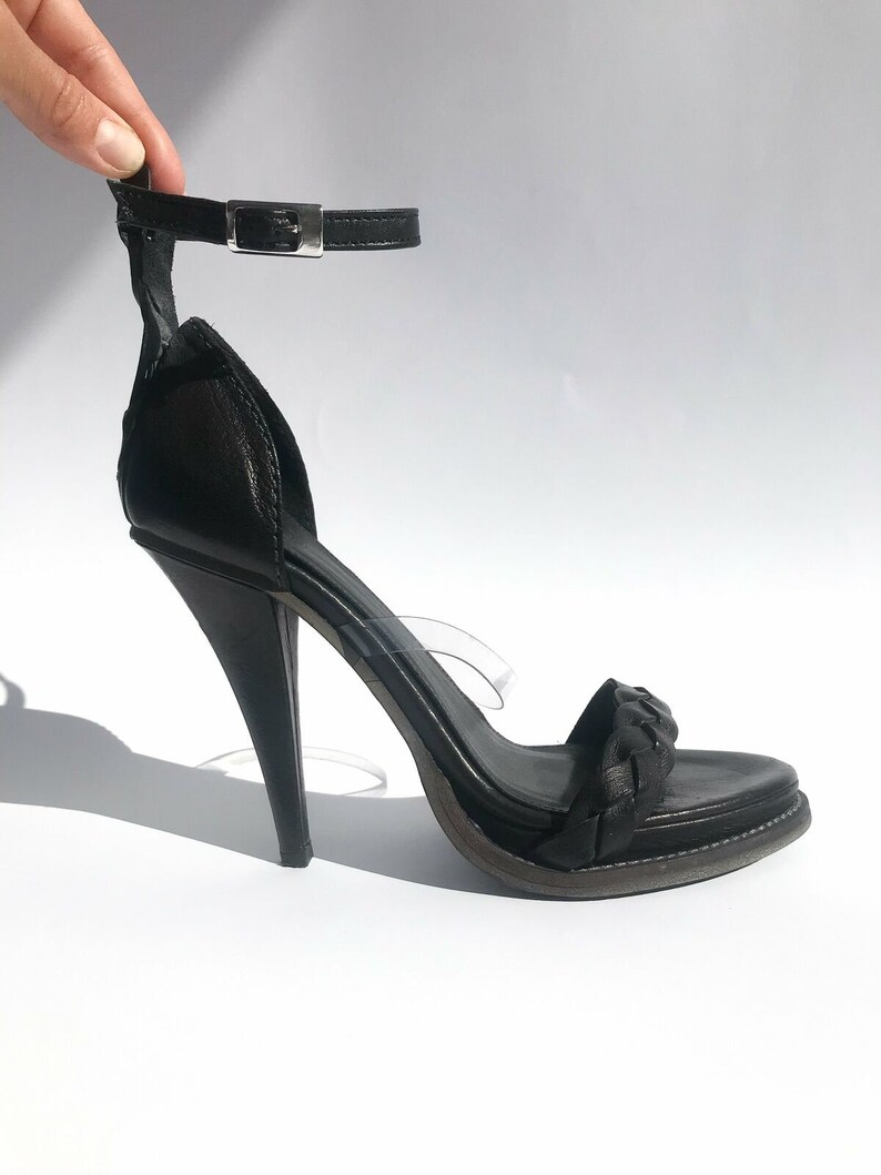 May include: Black leather heeled sandals with a braided strap and an ankle strap with a silver buckle.