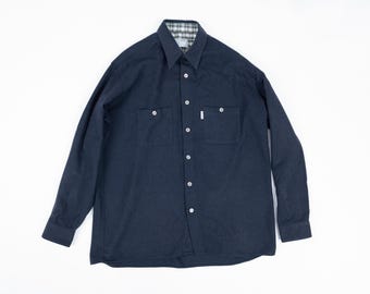 May include: A dark blue long-sleeved button-down shirt with a plaid lining on the collar. The shirt has two chest pockets and a button-up front.